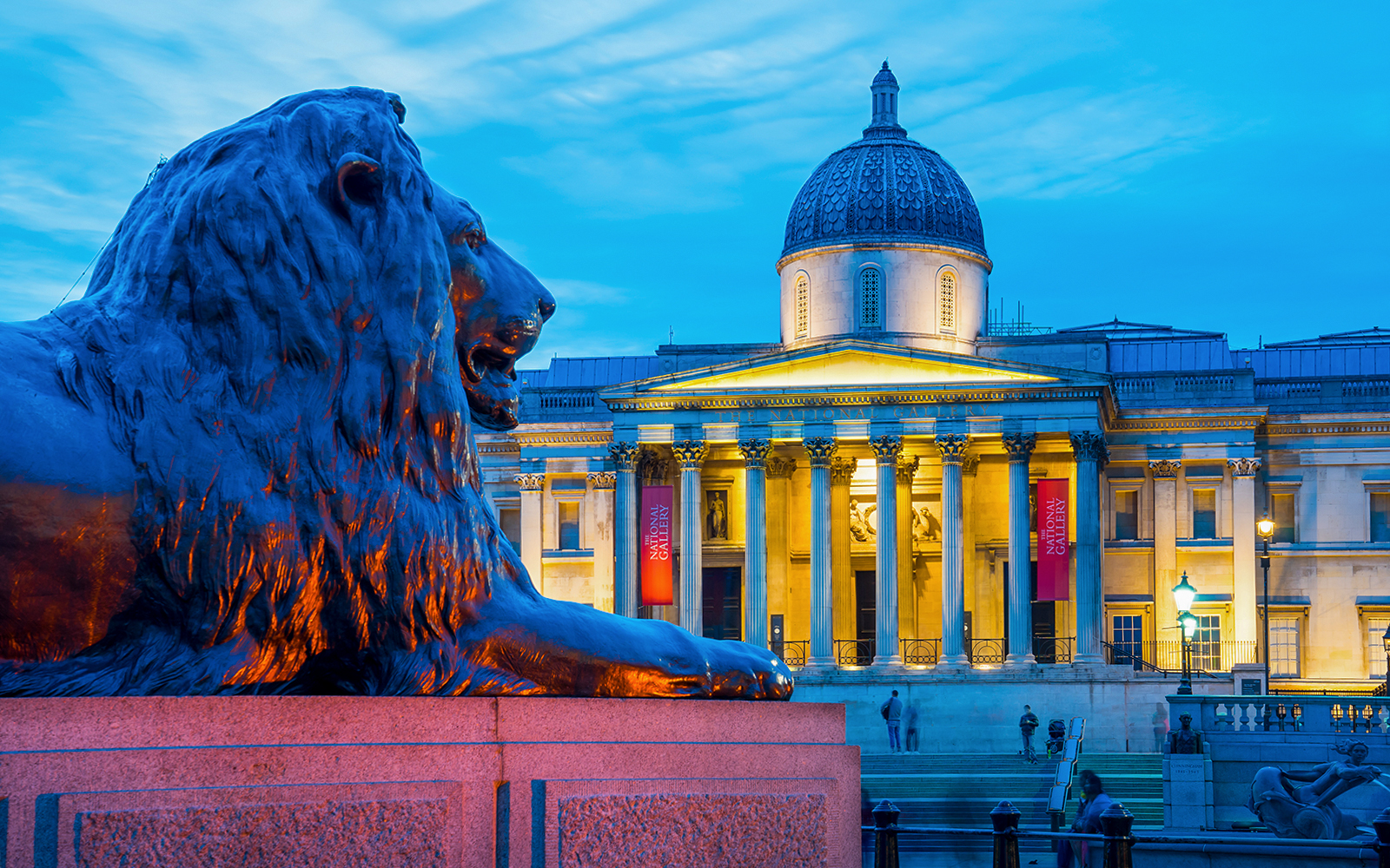 Trafalgar Square lion statue with the National Gallery in London at dusk.