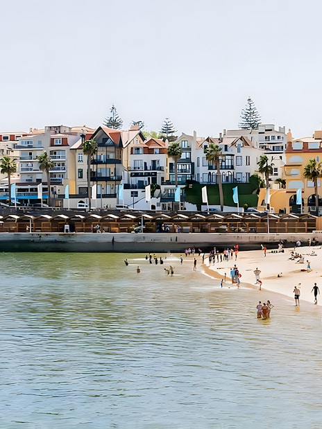 Beachfront in Cascais with people enjoying the sand and water, colorful buildings in the background.