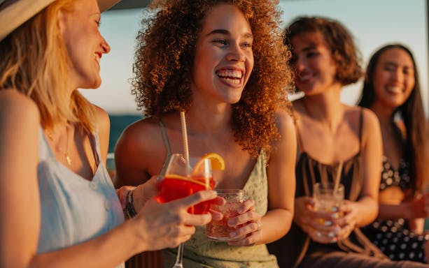 Friends laughing and holding cocktails at sunset during a boat party.