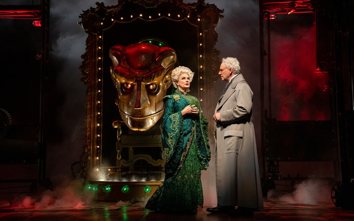 Stage scene from "Wicked" with two characters in elaborate costumes and a large mechanical face.