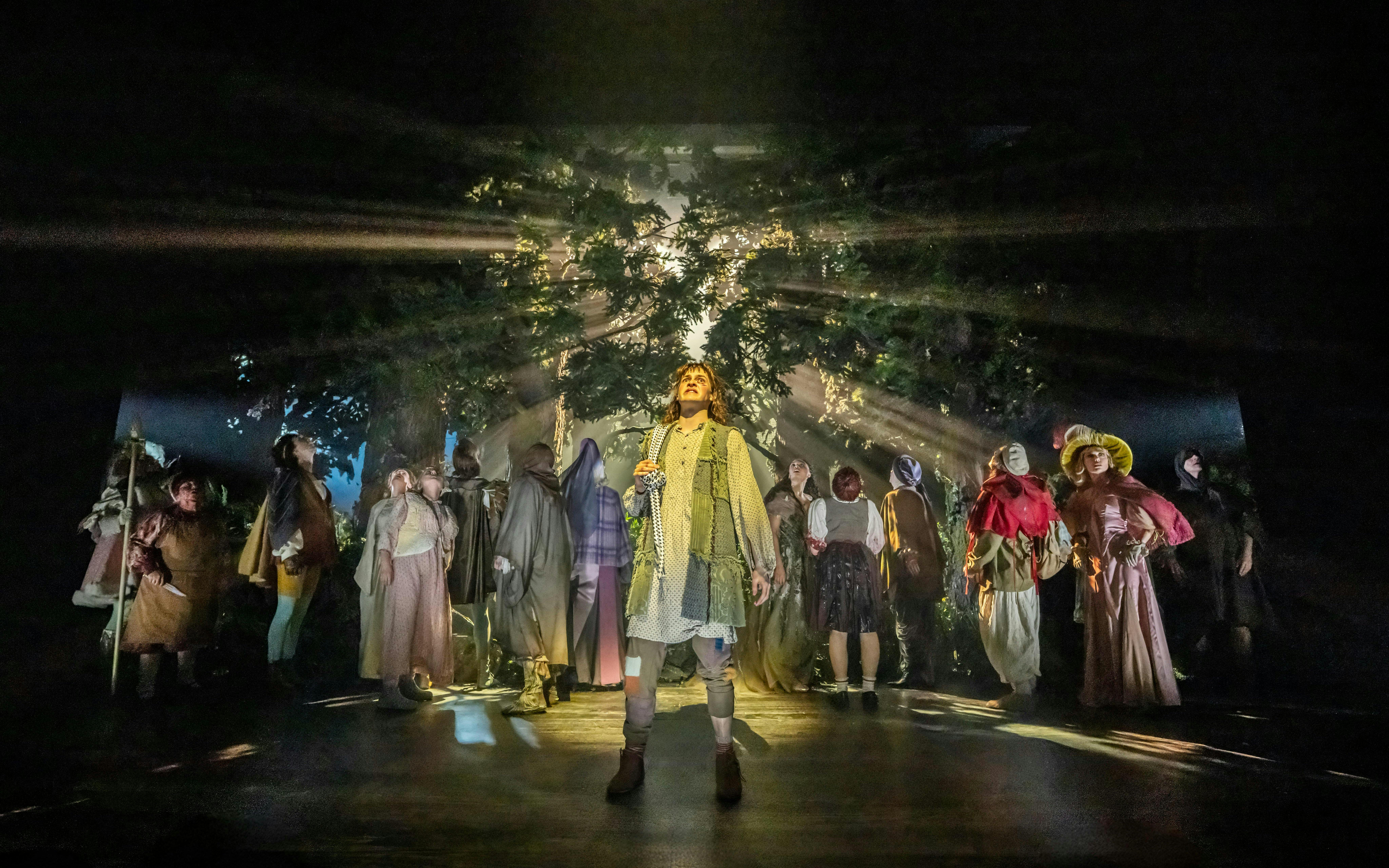 Cast performing in "Into The Woods" with forest backdrop and dramatic lighting.