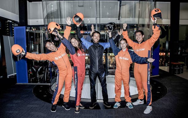 Indoor skydiving group with instructor at AirRider, wearing flight suits and holding helmets.