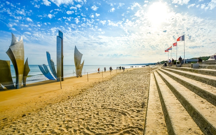 Tourists sit under flags on steps at Omaha Beach near Les Braves Memorial, Normandy.