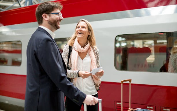 Travelers with luggage and tickets at a train station platform with a Eurail train.