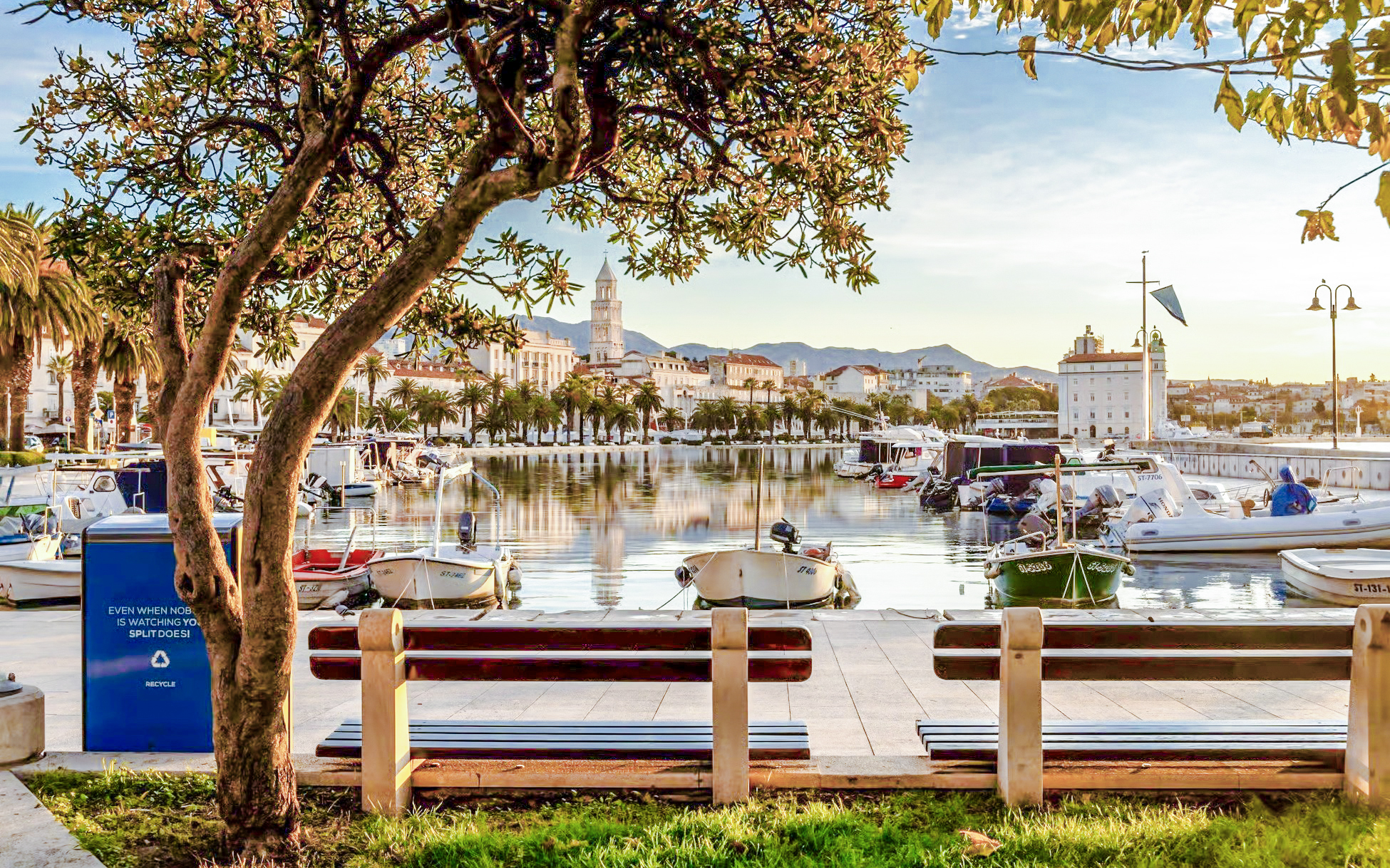 Boats docked along the Riva promenade in Split, Croatia with cityscape and mountains in the background.