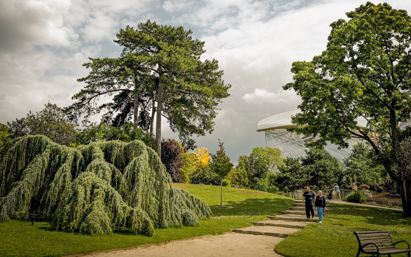 Visitors walking through Le Jardin d'Acclimatation with lush greenery and modern architecture in the background.