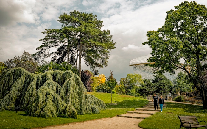 Visitors walking through Le Jardin d'Acclimatation with lush greenery and modern architecture in the background.