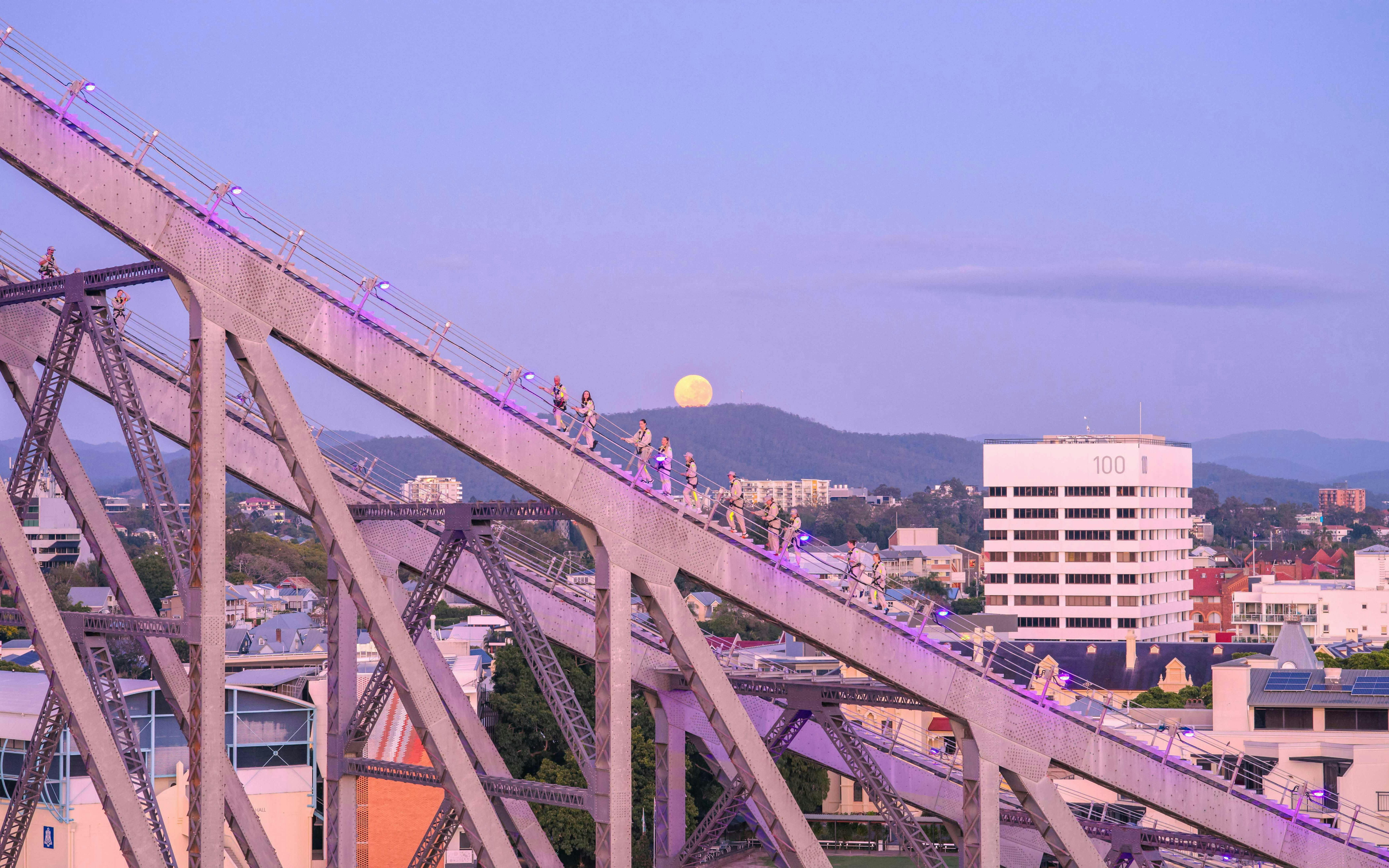 Climbers ascending Story Bridge in Brisbane at dusk with a full moon in the background.