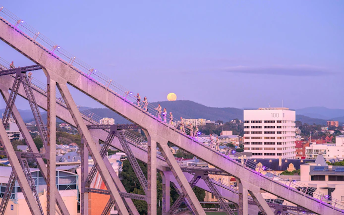 Climbers ascending Story Bridge in Brisbane at dusk with a full moon in the background.
