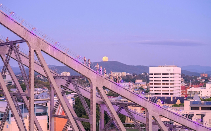 Climbers ascending Story Bridge in Brisbane at dusk with a full moon in the background.