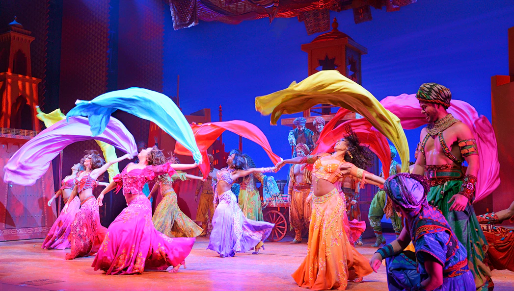 Performers in colorful costumes dancing with flowing scarves in an Aladdin stage show.