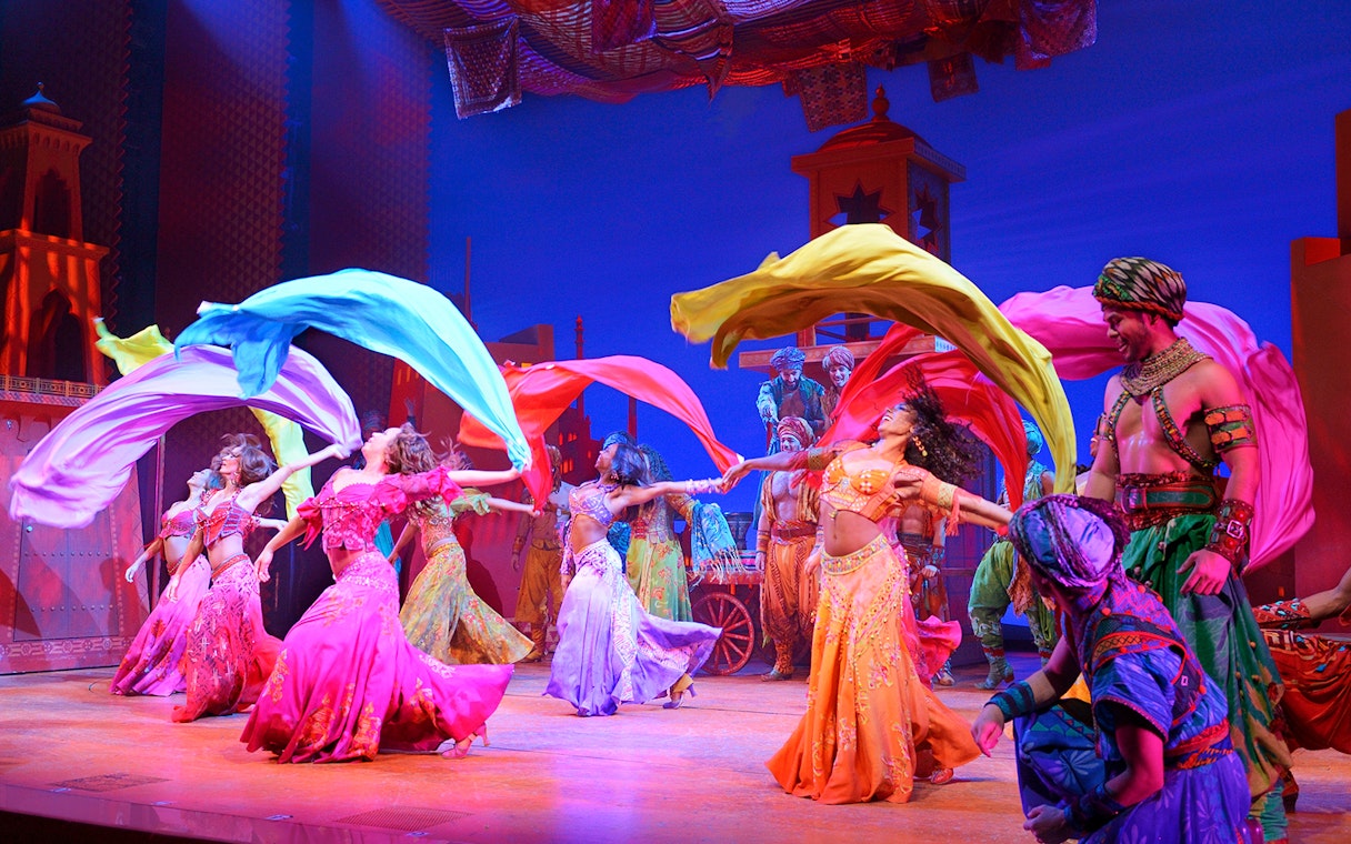 Performers in colorful costumes dancing with flowing scarves in an Aladdin stage show.