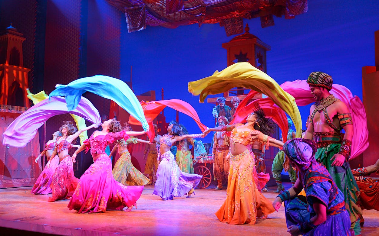 Performers in colorful costumes dancing with flowing scarves in an Aladdin stage show.