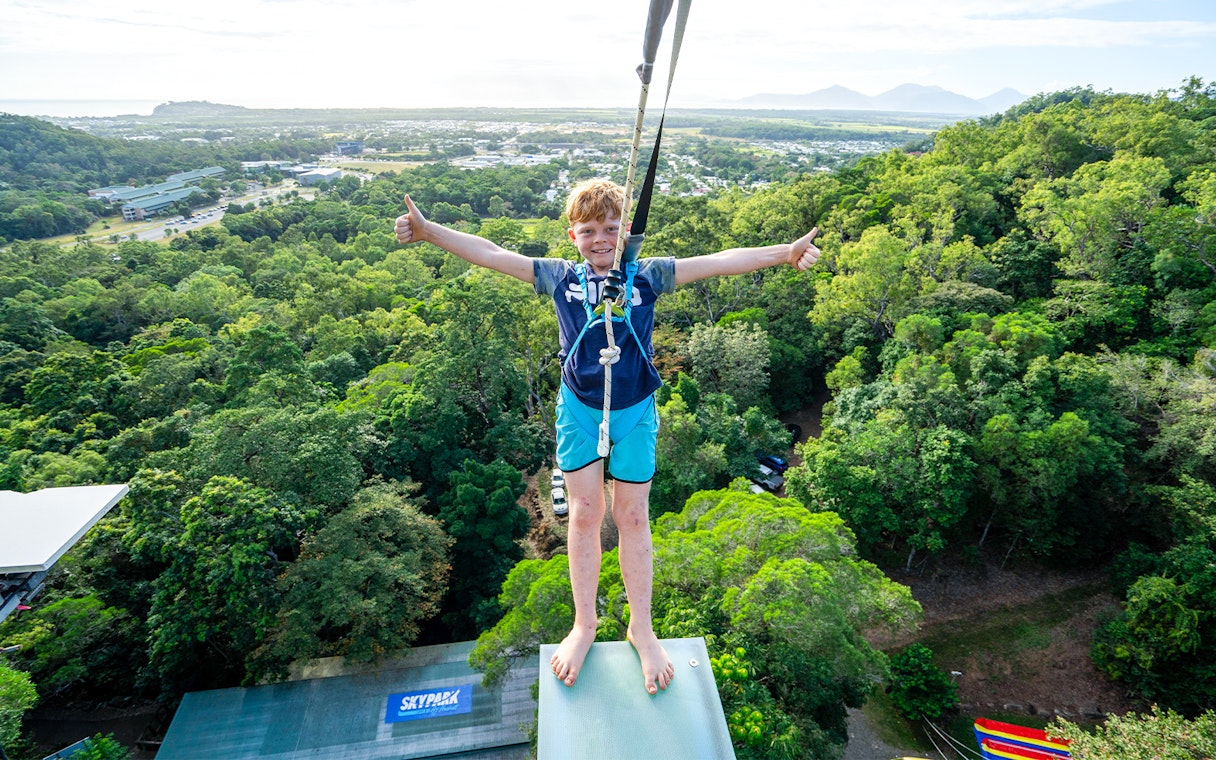 Young person harnessed on Walk The Plank by AJ Hackett, overlooking lush forest and distant mountains.