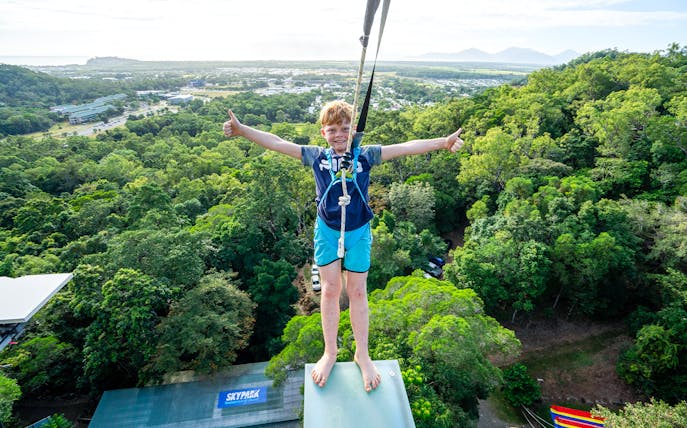 Young person harnessed on Walk The Plank by AJ Hackett, overlooking lush forest and distant mountains.
