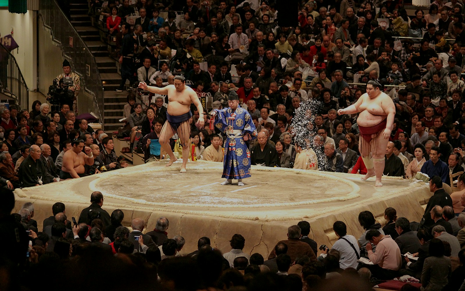 Sumo wrestlers and referee in a tournament ring, Osaka audience watching.