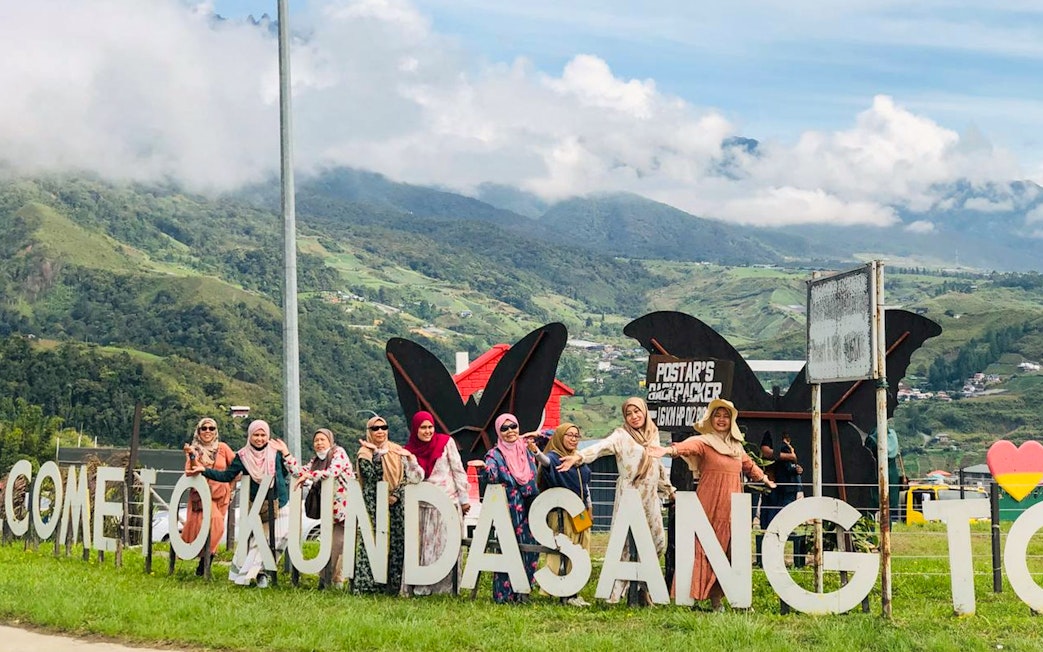 Group posing by "Welcome to Kundasang" sign with Mount Kinabalu in the background.