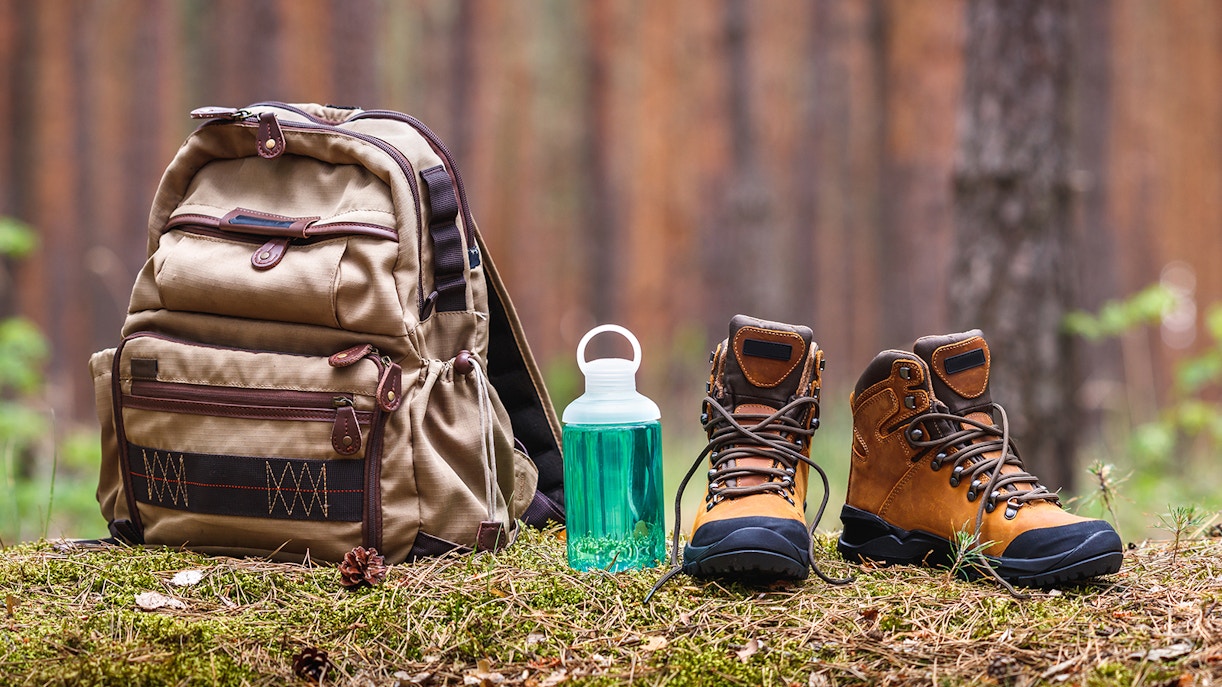 Backpack, water bottle, and leather boots on forest floor for hiking and camping.