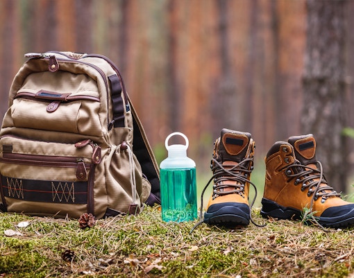 Backpack, water bottle, and leather boots on forest floor for hiking and camping.