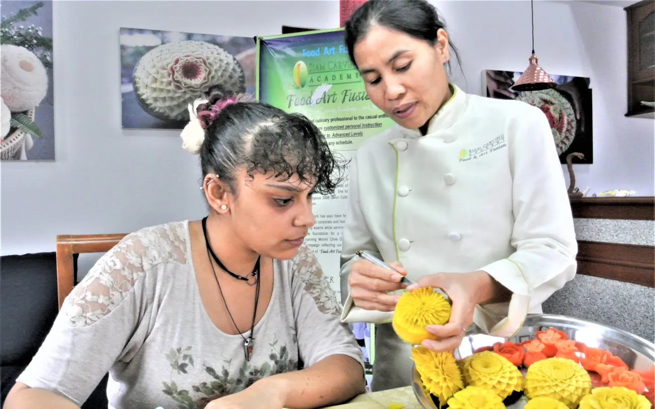 Thai master carver teaching fruit carving to a student in a class.