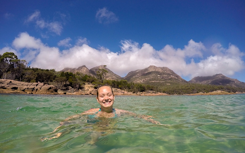 Person swimming in clear water at Wineglass Bay with mountain backdrop.