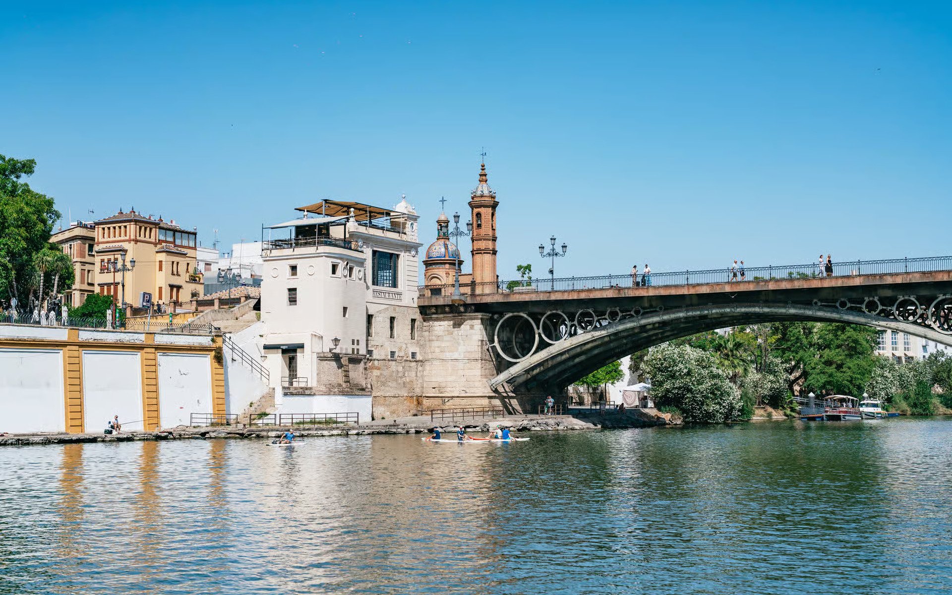 Triana Bridge over Guadalquivir River in Seville, Spain, with people walking and kayaking below.