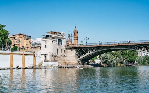 Triana Bridge over Guadalquivir River in Seville, Spain, with people walking and kayaking below.