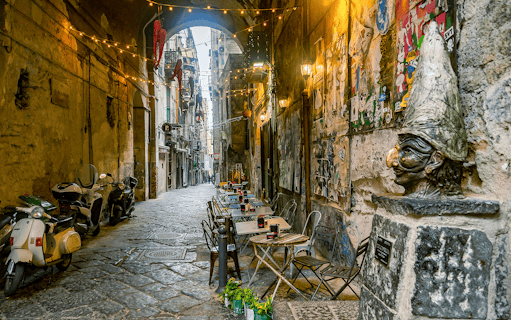 Narrow street in Naples with outdoor café tables and a Pulcinella statue.