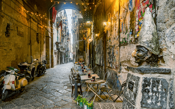 Narrow street in Naples with outdoor café tables and a Pulcinella statue.
