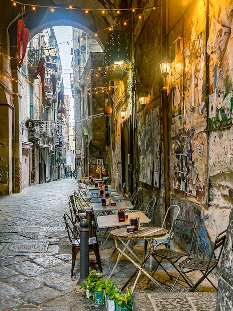 Narrow street in Naples with outdoor café tables and a Pulcinella statue.