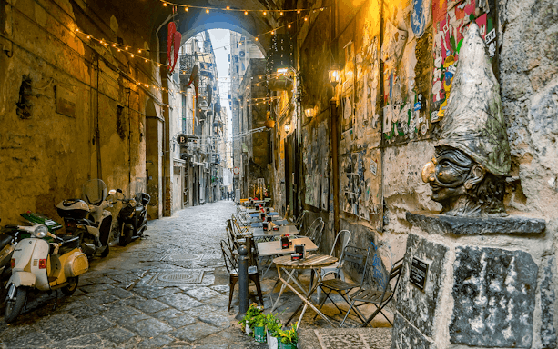 Narrow street in Naples with outdoor café tables and a Pulcinella statue.