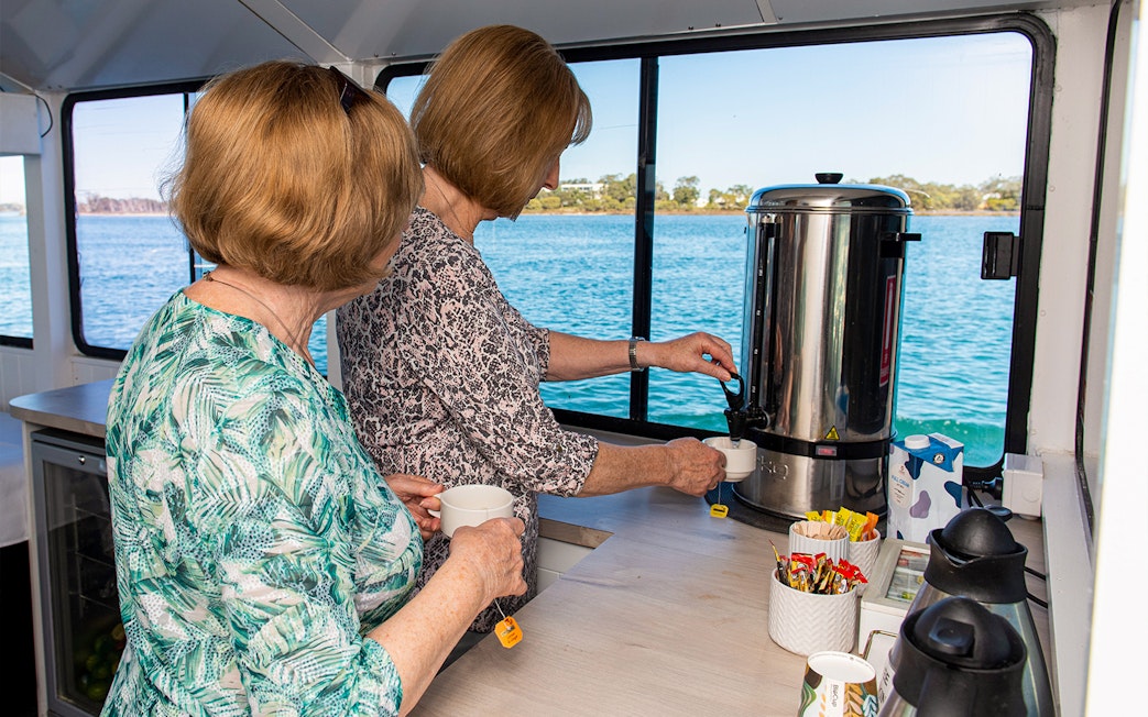 Two people preparing tea on a Murray River lunch cruise with a scenic water view.