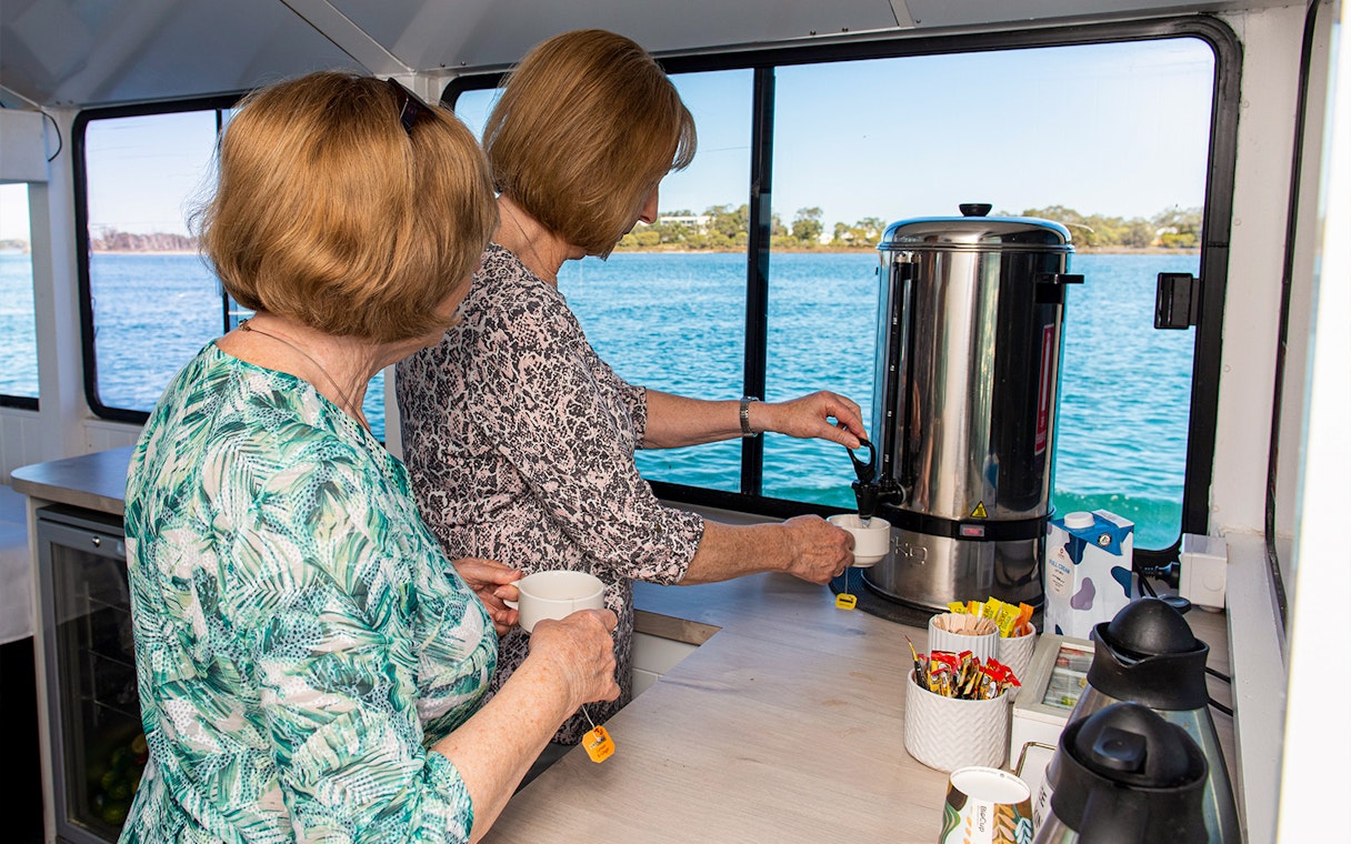 Two people preparing tea on a Murray River lunch cruise with a scenic water view.