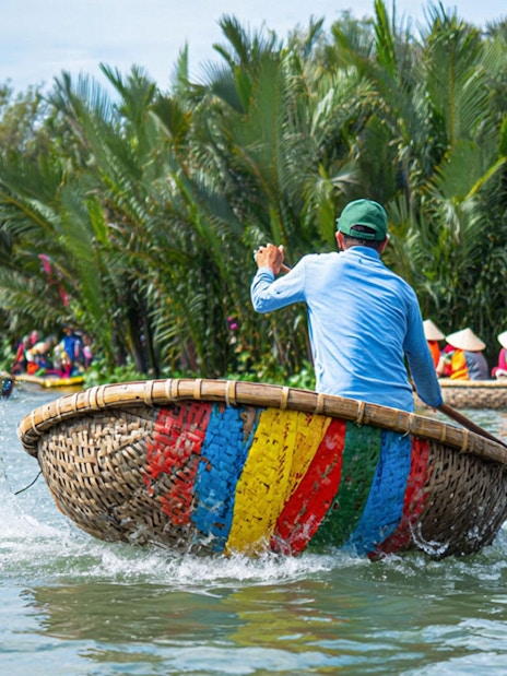 Guide rowing a colorful basket boat on Hoi An river with tourists in the background.