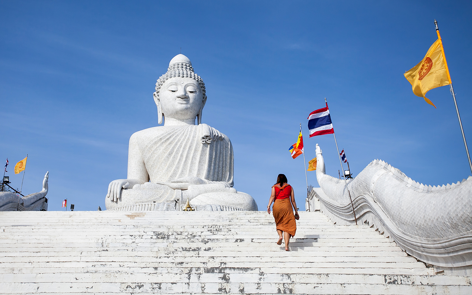 Woman ascending steps to Big Buddha statue in Phuket, Thailand, with flags in the background.