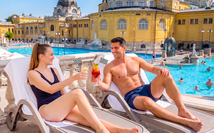 Couple enjoying cocktails at Szechenyi Spa, Budapest, with pool and historic architecture in view.