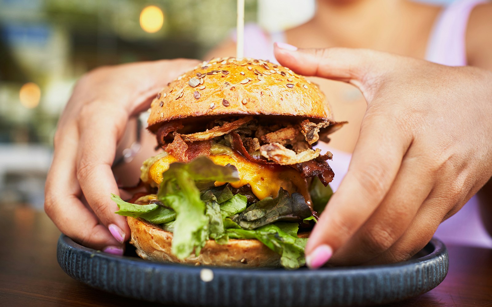 Hands holding a juicy hamburger with lettuce, cheese, and bacon on a seeded bun.