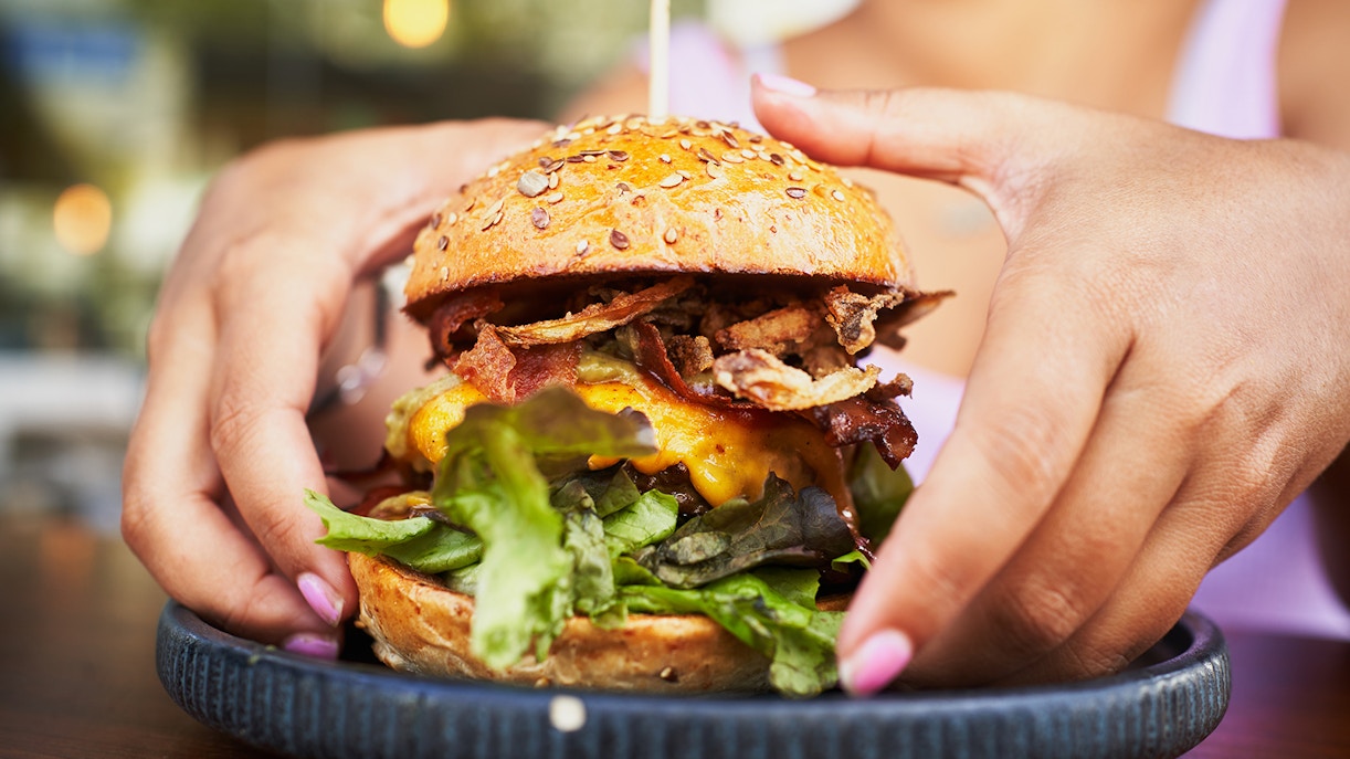 Hands holding a juicy hamburger with lettuce, cheese, and bacon on a seeded bun.