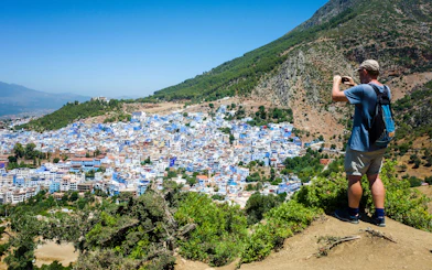 Traveler capturing panoramic view of Chefchaouen, Morocco, with blue buildings on Rif mountain slopes.