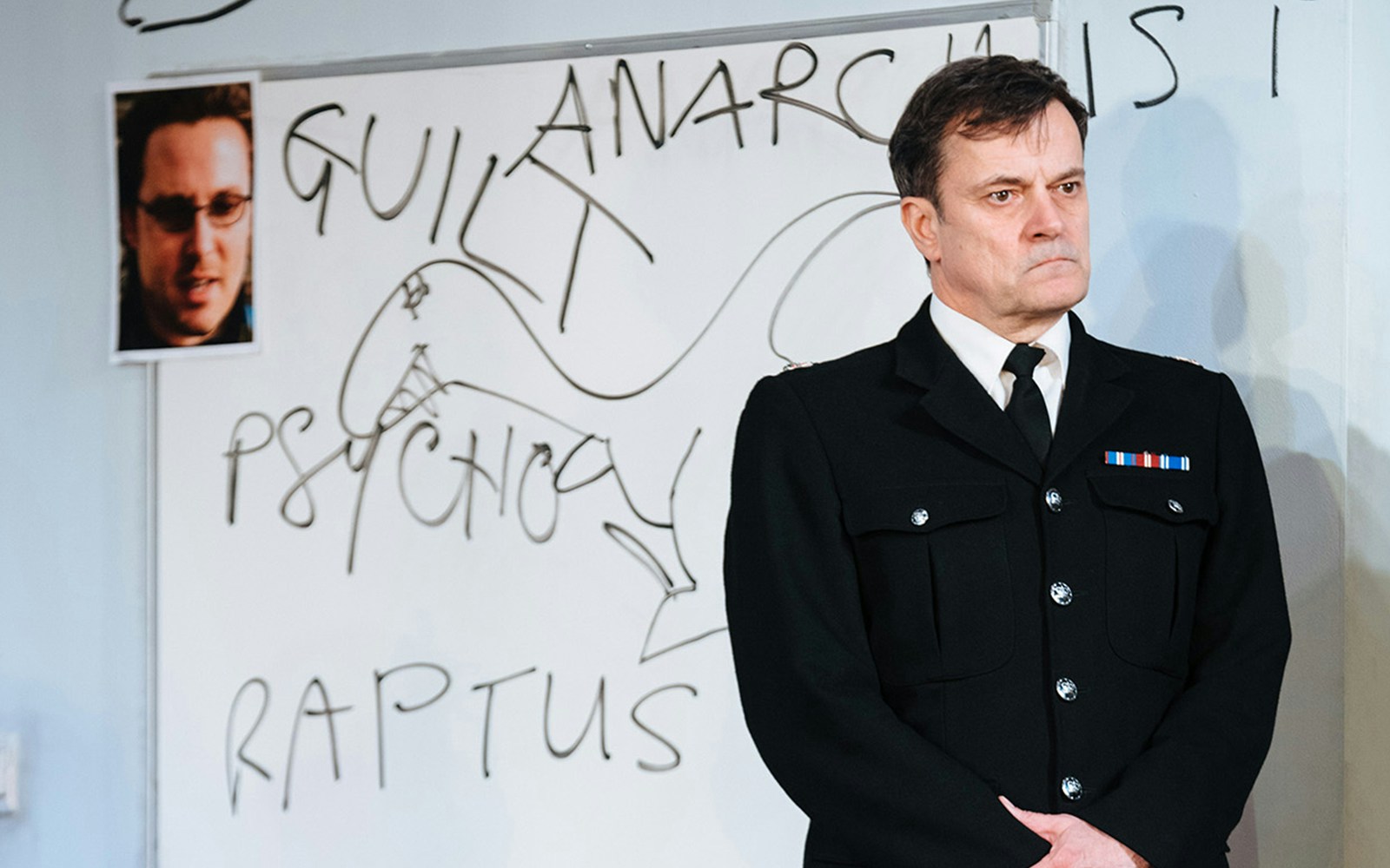 Police officer stands in front of a whiteboard with words related to "Accidental Death of an Anarchist" play.