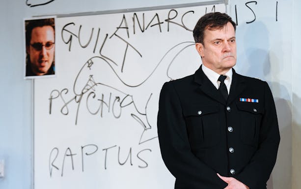 Police officer stands in front of a whiteboard with words related to "Accidental Death of an Anarchist" play.