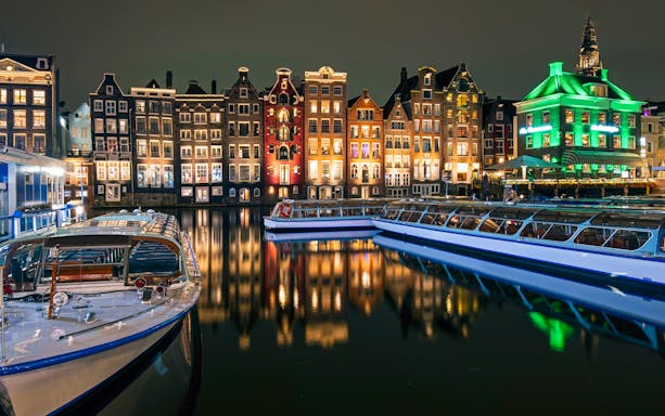 Amsterdam canal boats with illuminated historic buildings at night.