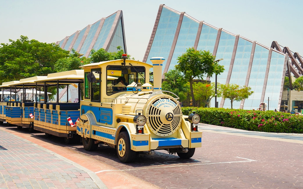 Dubai Safari Park shuttle train in front of modern glass building.
