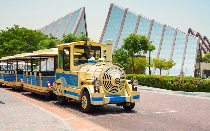 Dubai Safari Park shuttle train in front of modern glass building.