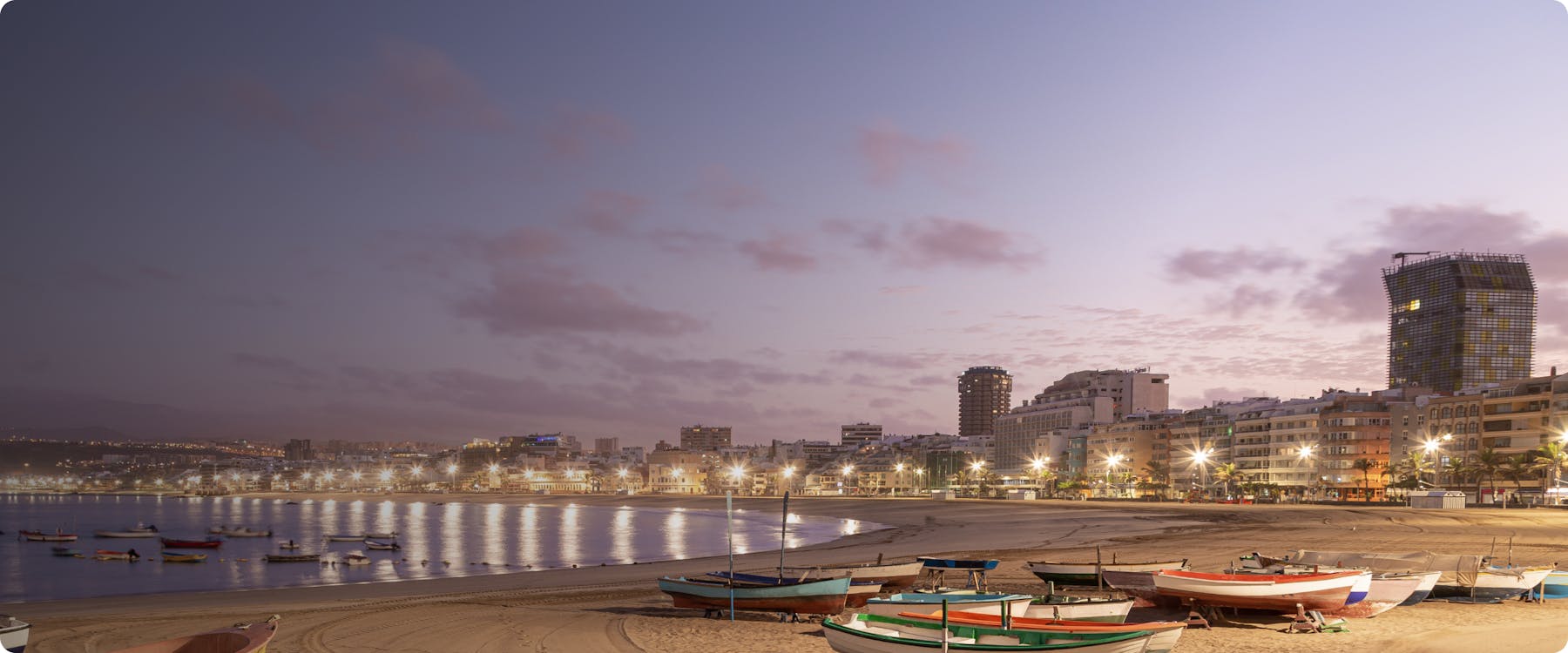 Las Palmas de Gran Canaria beach at dusk with boats and city skyline.