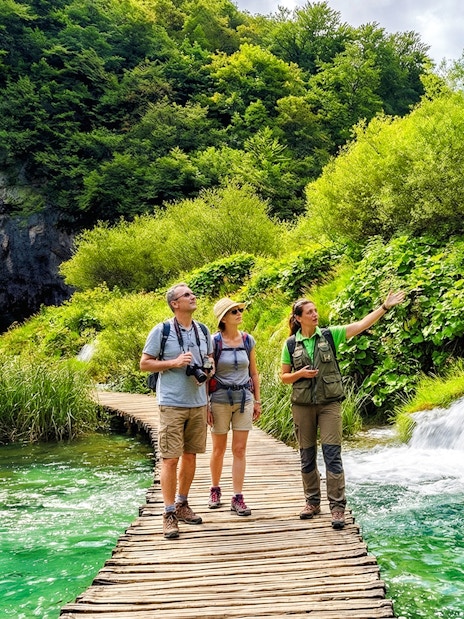 Guests with a tour guide on a wooden path by waterfalls in Plitvice Lakes National Park.