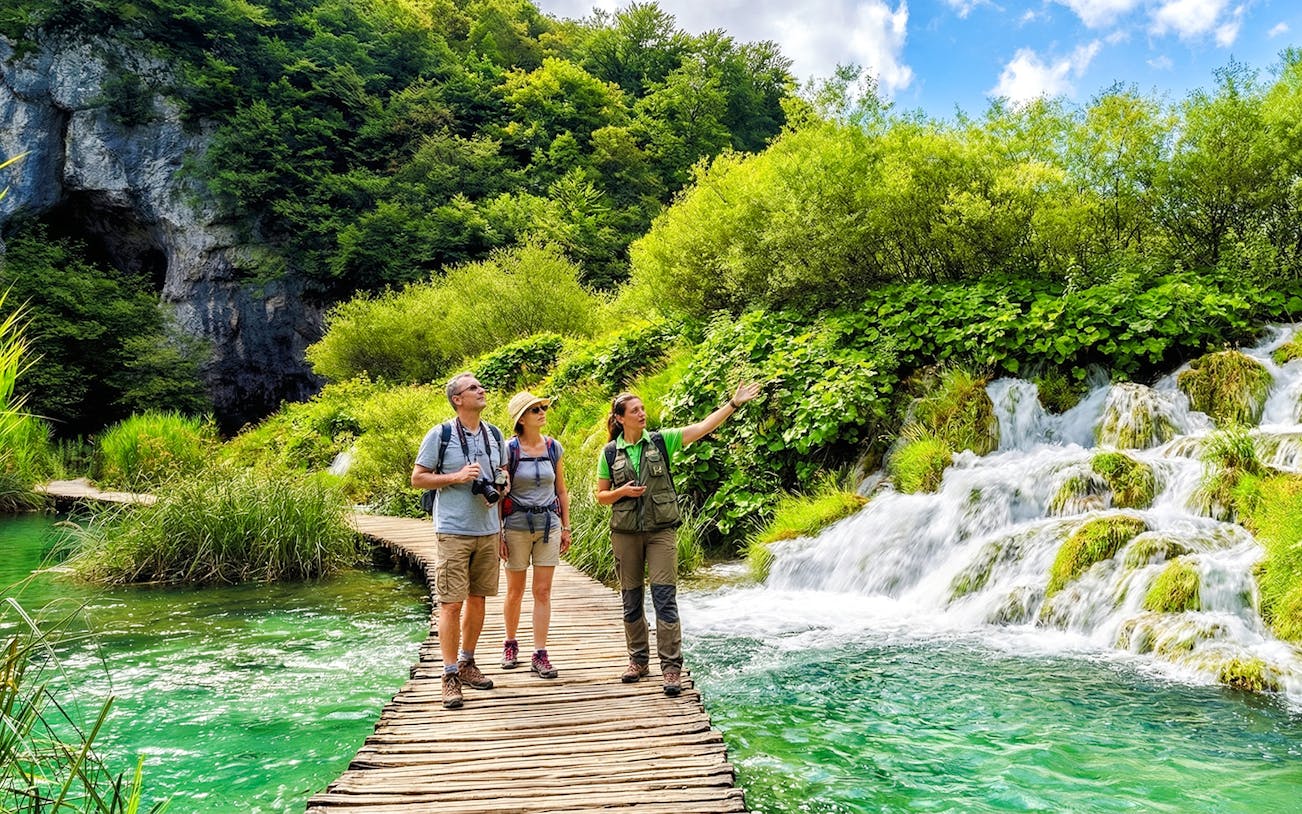 Guests with a tour guide on a wooden path by waterfalls in Plitvice Lakes National Park.