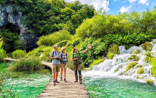Guests with a tour guide on a wooden path by waterfalls in Plitvice Lakes National Park.