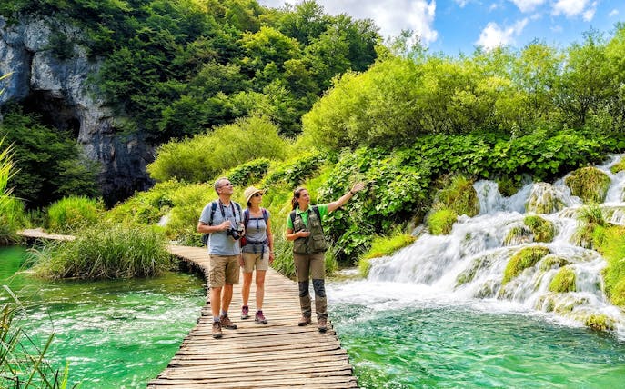 Guests with a tour guide on a wooden path by waterfalls in Plitvice Lakes National Park.