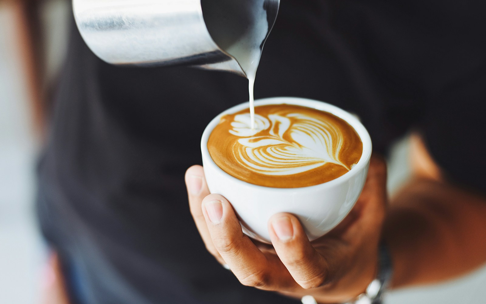 Barista pouring milk into a latte with art design in a white cup.